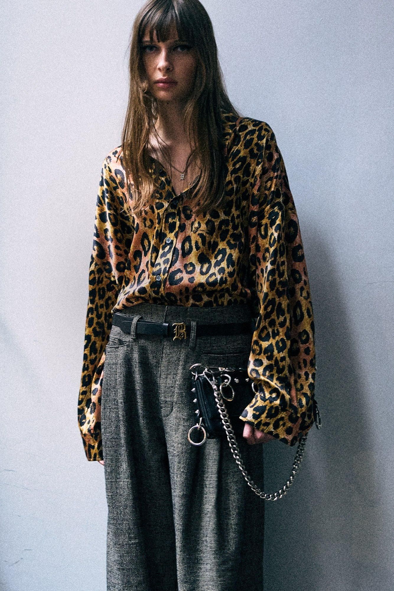 A model wearing a long-sleeve leopard print blouse with a black belt and dark gray wide-leg trousers, standing against a textured gray wall.