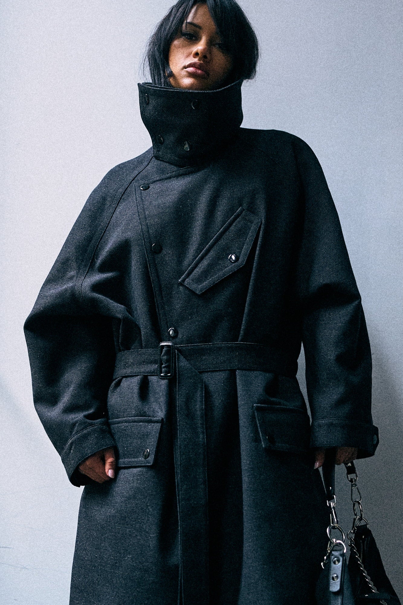 A model wearing a dark charcoal gray high-neck coat with a belted waist, shown from the head down against a plain light gray background.