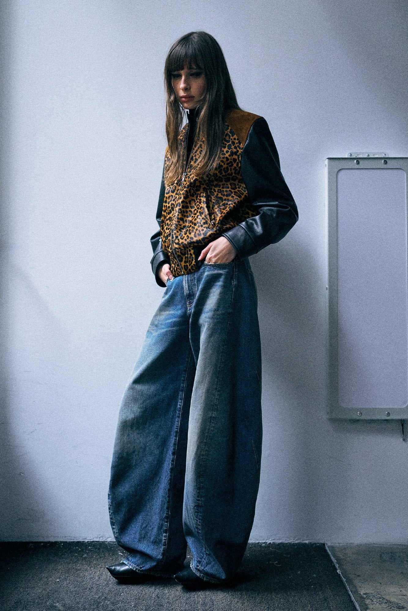 A model stands against a light gray wall wearing a black long-sleeve top with a brown and black leopard-print vest, paired with very wide-leg blue jeans.