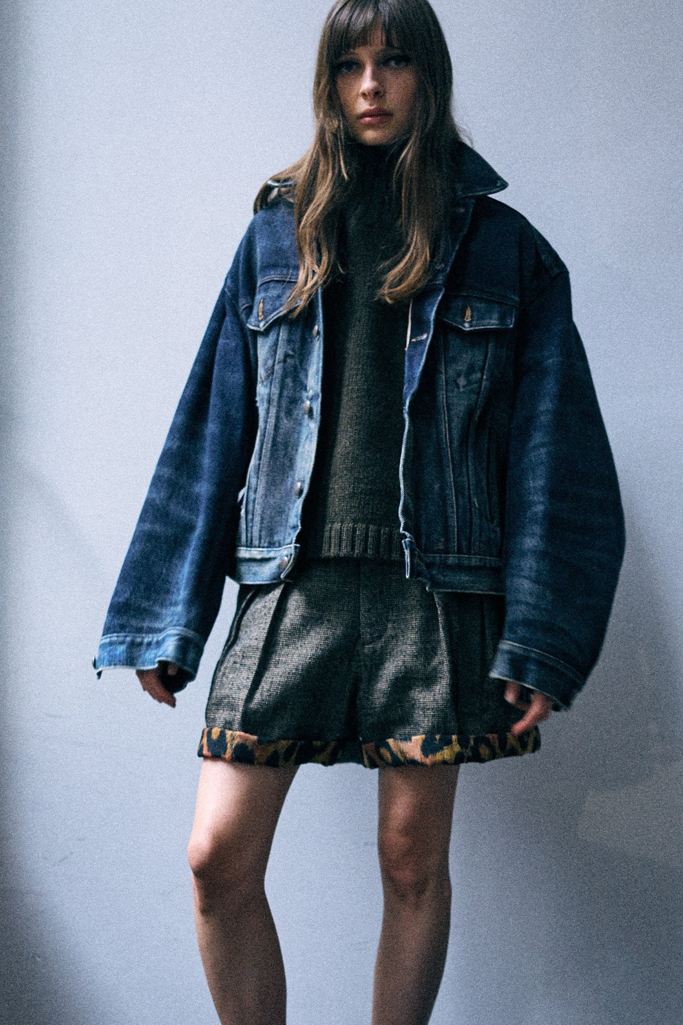 A model wearing a dark blue oversized denim jacket over a dark gray knit sweater and matching dark gray pleated shorts with a patterned hem, standing against a plain gray wall.