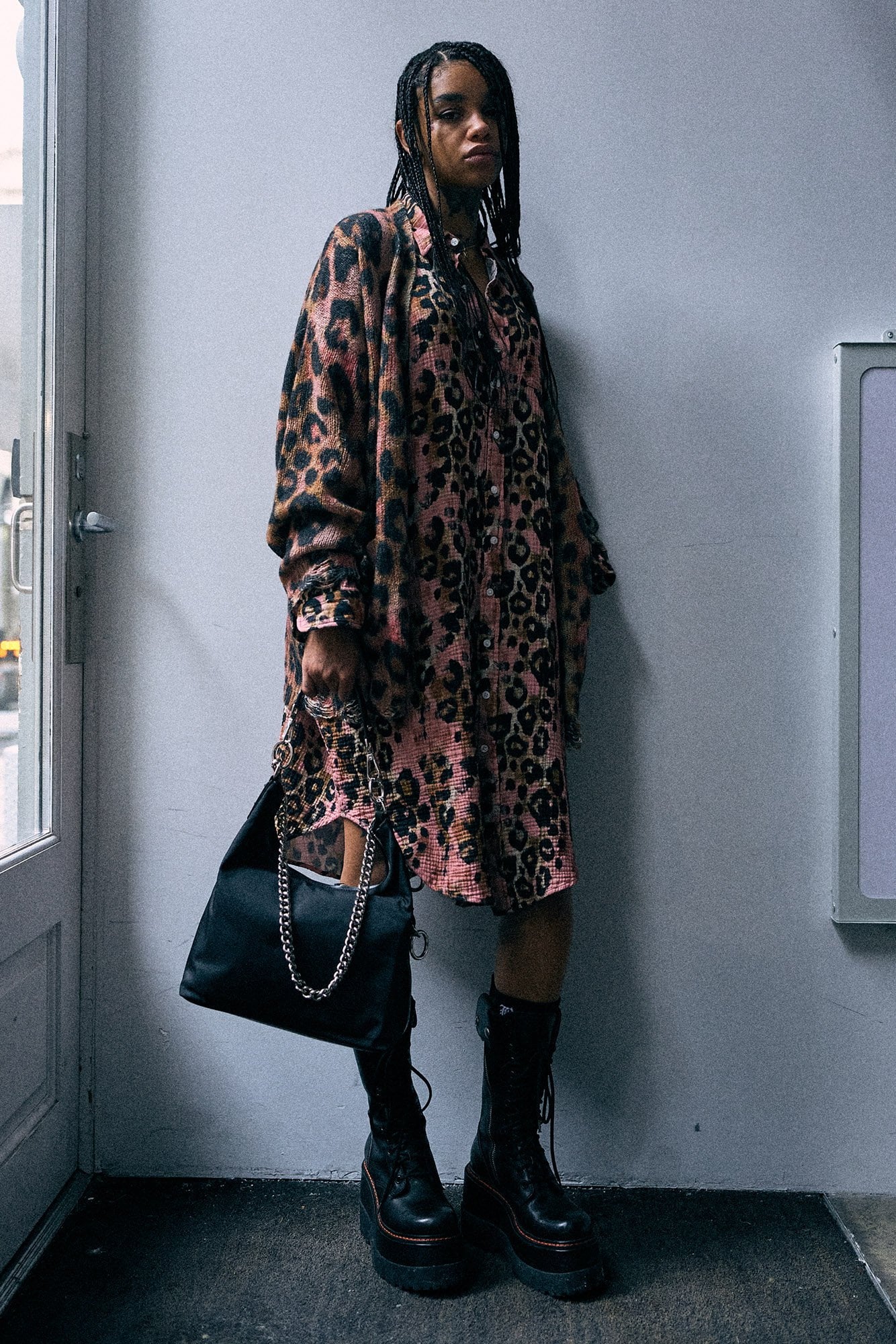 A model stands in a corner wearing an oversized pink leopard-print coat over a matching top, paired with black combat boots and carrying a large black shoulder bag.