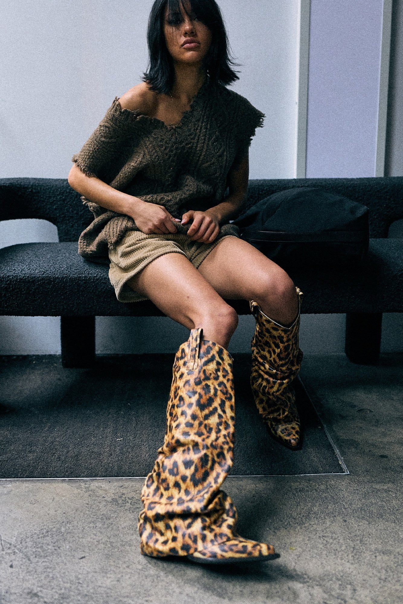 A model sits on a dark bench wearing a brown knit off-the-shoulder top, beige shorts, and a pair of leopard-print slouchy knee-high boots in a minimalist indoor setting.
