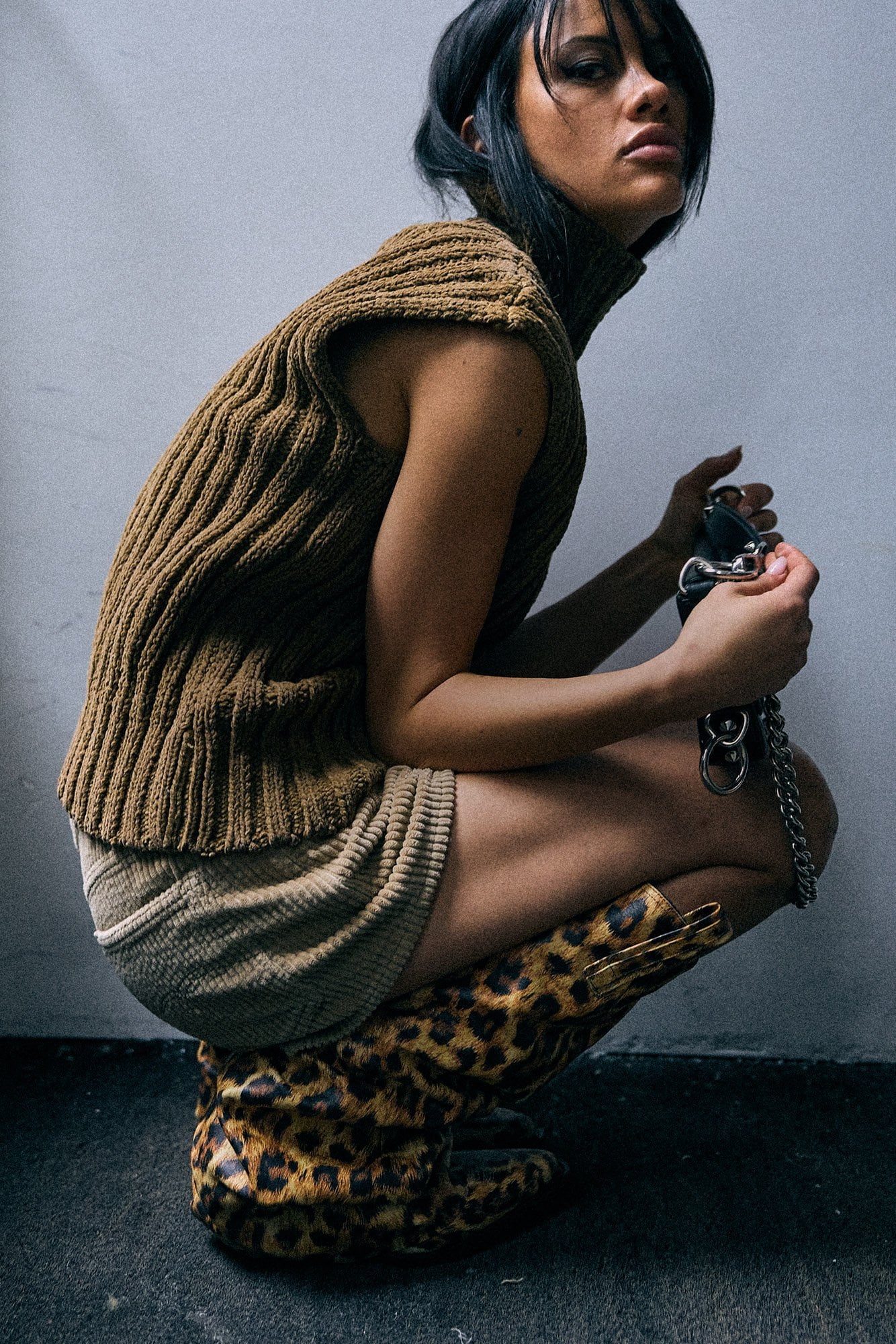 A model crouches against a gray wall wearing a brown ribbed knit sleeveless top, a beige knit skirt, and leopard-print boots, holding a small black handbag with a chain strap.