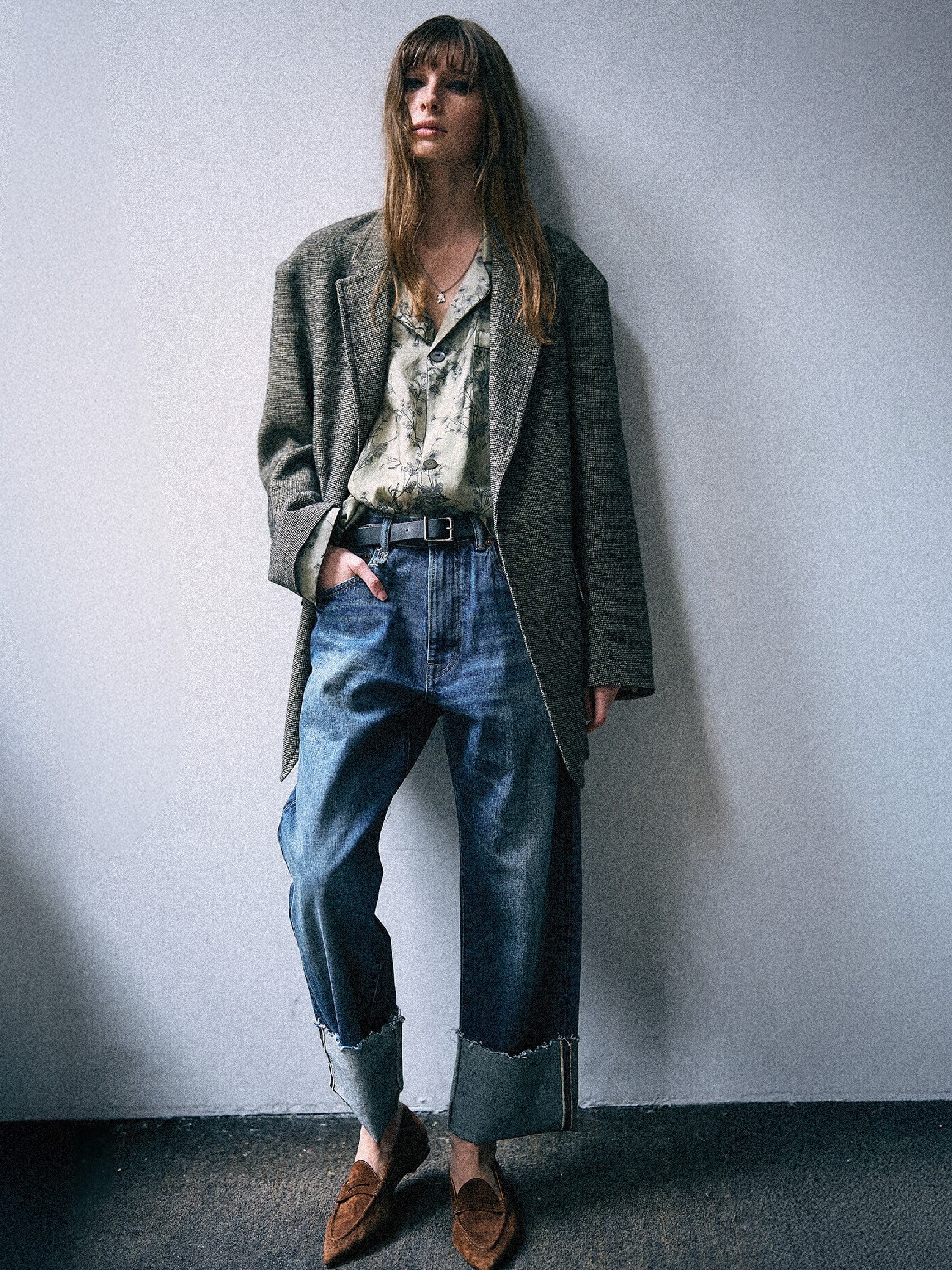 A model stands against a plain light gray wall wearing a long gray textured blazer over a light patterned shirt, blue jeans with rolled cuffs, and brown loafers.