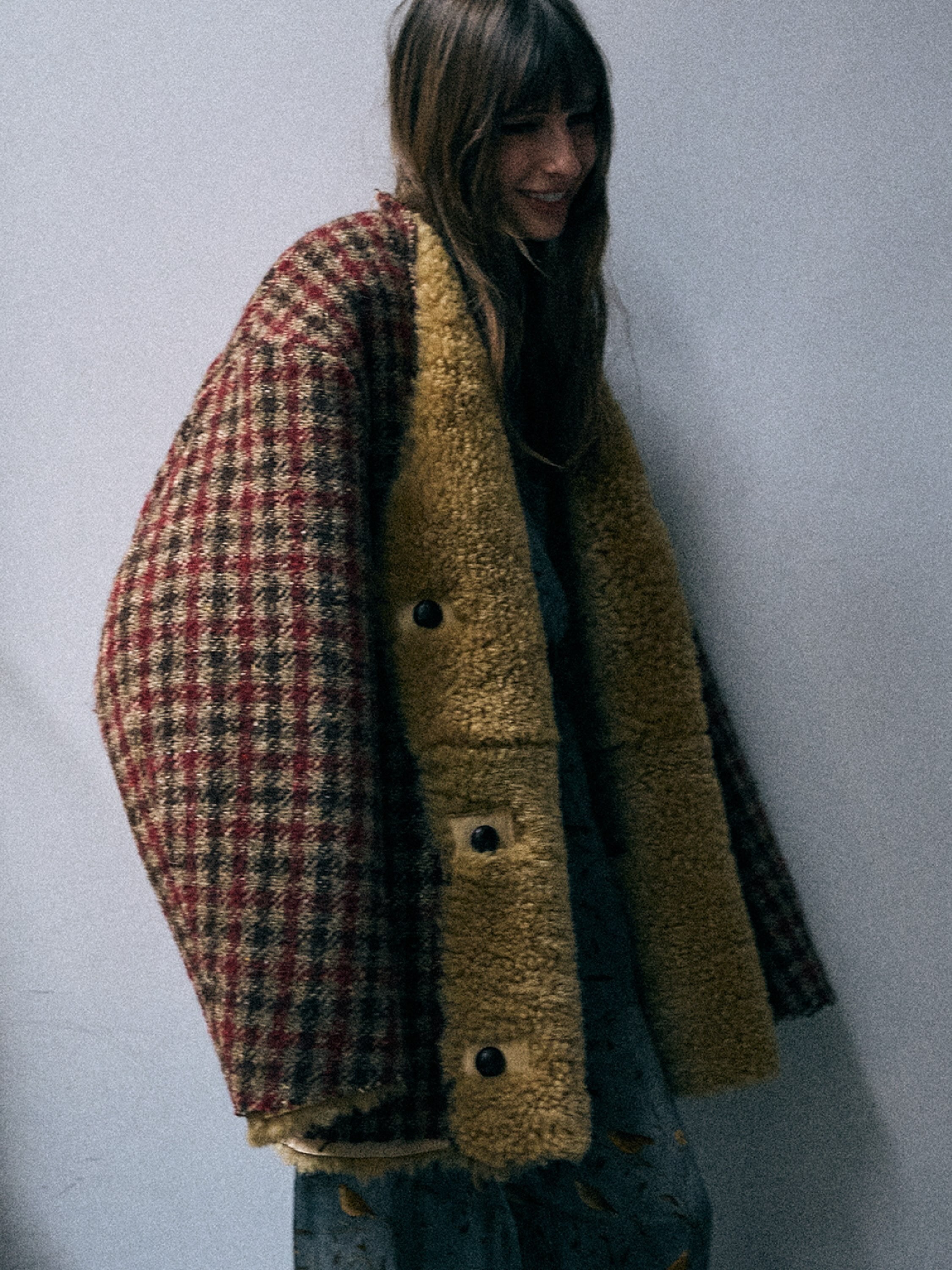 A person wearing a layered outerwear piece with a red, brown, and black plaid wool panel on one side and a tan sherpa fleece panel on the other, styled over blue floral pj pants.