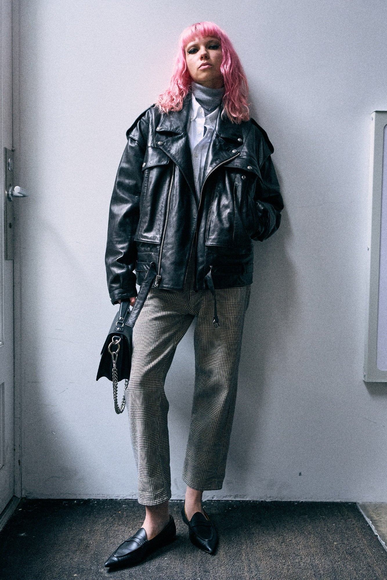 A person with bright pink hair stands against a plain light gray wall wearing an oversized black leather jacket over a white top, paired with loose cropped trousers and black pointed loafers, while holding a black handbag.