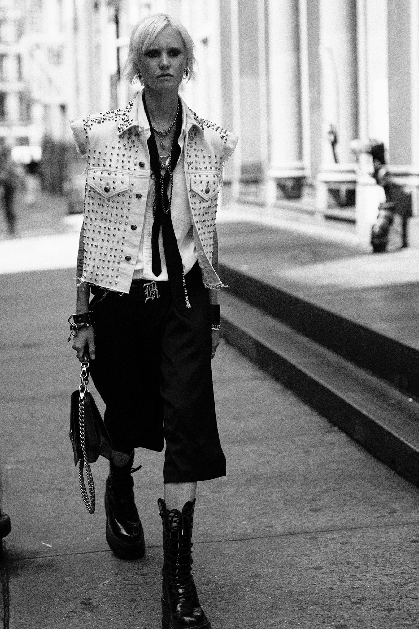 Model walks down the sidewalk wearing a studded white trucker vest and black shorts.