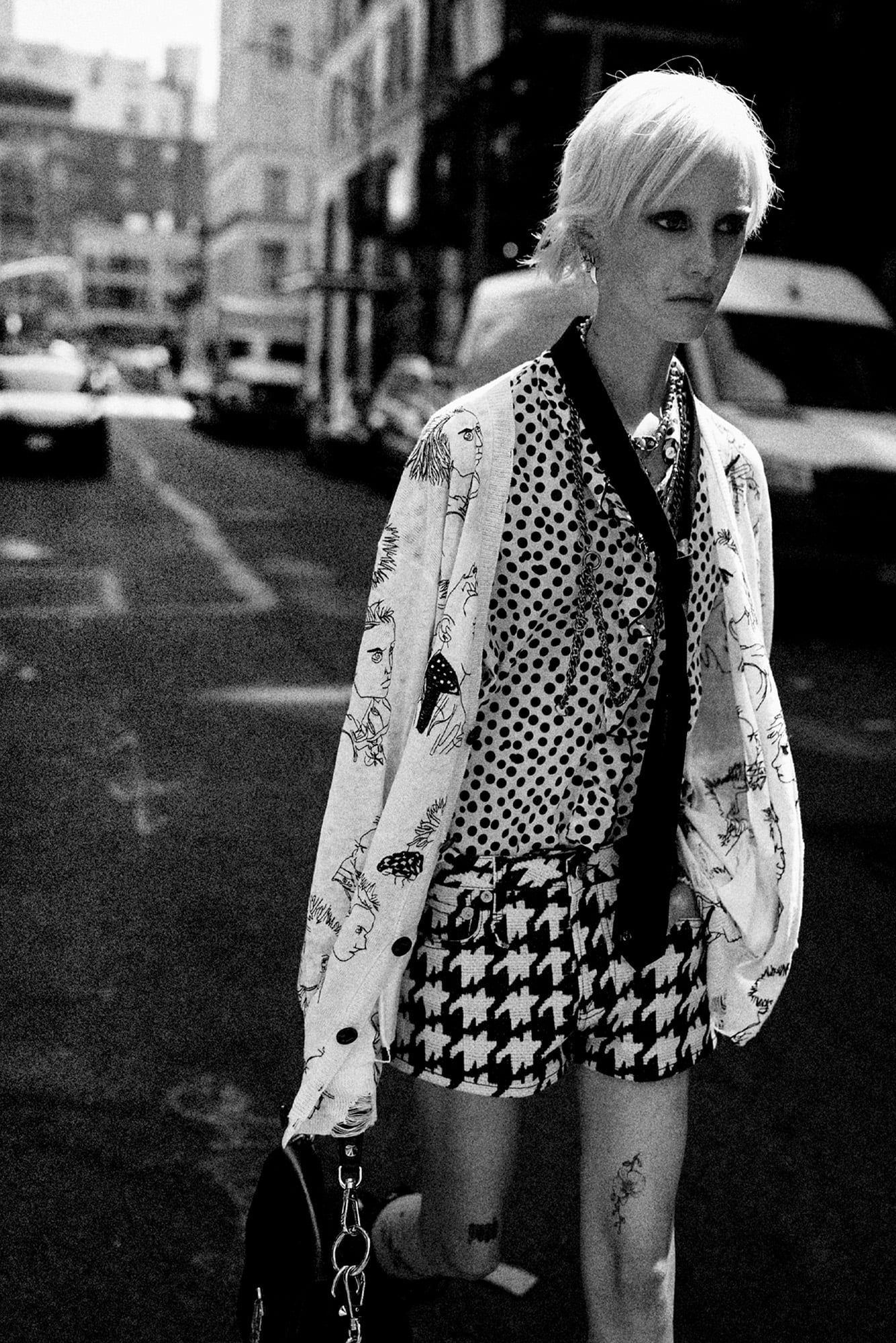 Model walking down the street wearing a patterned cardigan and blouse with black and white houndstooth shorts.